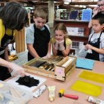 An adult demonstrates the art of letterpress to a group of four kids.