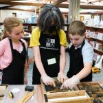 Three kids learn as an adult shows them how to use a letterpress.