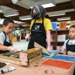 An adult shows two kids how to use a letterpress kit.